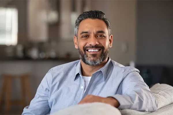 BioTE-Testosterone-Pellet-Clinic A mature South Asian man seated comfortably on a couch, smiling and looking directly at the camera satisfied with BioTE Testosterone Pellet Therapy offered by Anita Petruzzelli, M.D. in Glastonbury.