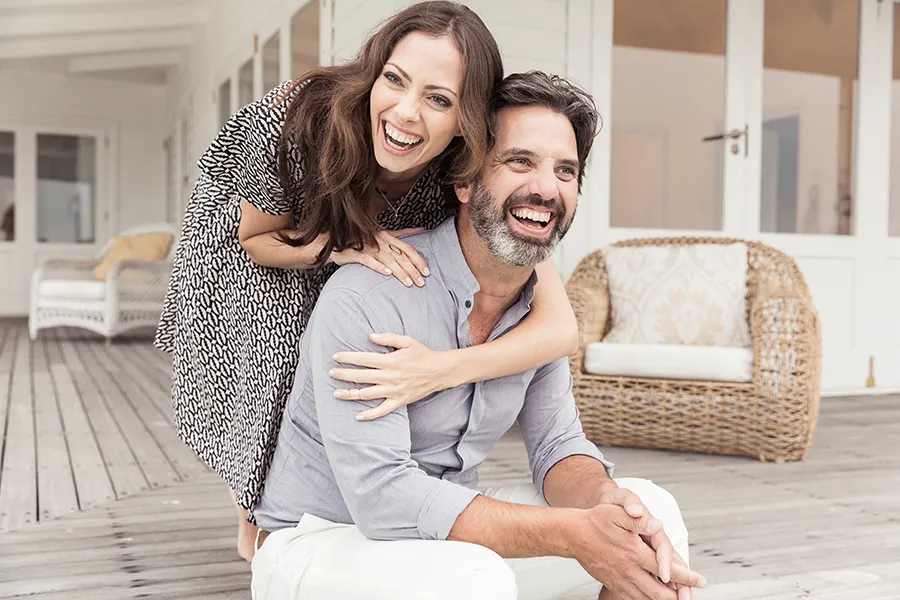 Health-and-Wellness-Services-Doctor A middle-aged couple on a wooden porch with wicker furniture; the woman is playfully hugging the man from behind. They are benefiting from the health and wellness services from Anita Petruzzelli, M.D. in Glastonbury.