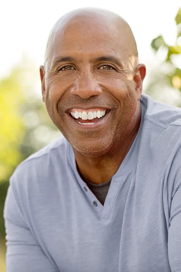 Healthy-Aging-Peptides-Treatment A middle-aged black man in a gray v-neck t-shirt sits outside smiling, happy with the healthy aging peptide treatments from Anita Petruzzelli, M.D. in Glastonbury