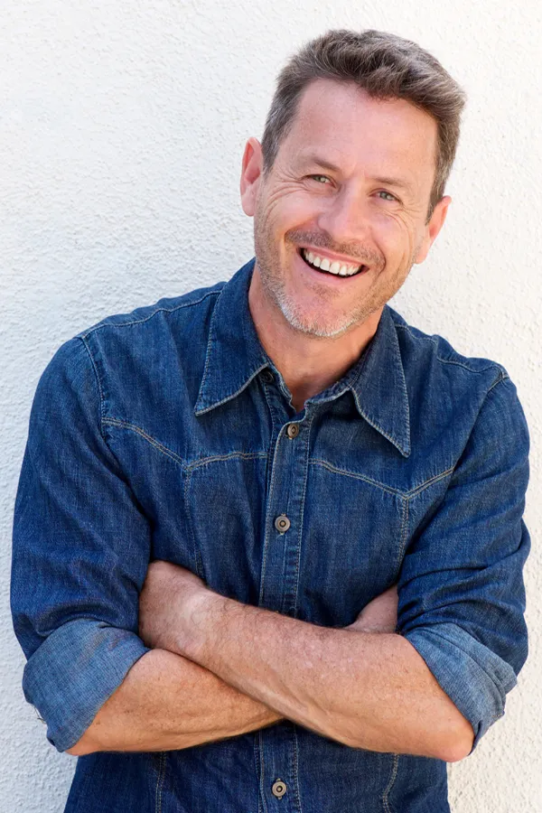 Ipamorelin-Peptide-Treatment A healthy middle-aged man in a denim shirt leans against a white wall smiling. Get Ipamorelin peptide treatment from Anita Petruzzelli, M.D. in Glastonbury.