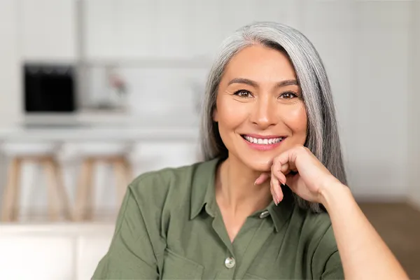 Menopause-Clinic An asian woman with long gray hair and a green shirt sitting and smiling on her couch, happy with her menopause care from [NAME} in Glastonbury.