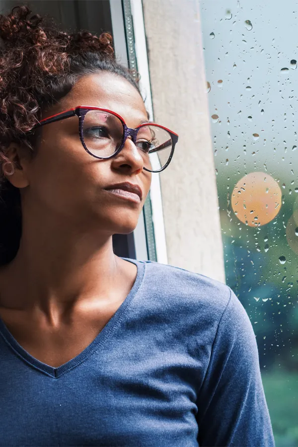 PMDD-Treatment A black woman with with a somber expression and glasses and a gray shirt stares out the window on a rainy day. Get treatment for Premenstrual Dysphoric Disorder from Anita Petruzzelli, M.D. in Glastonbury.