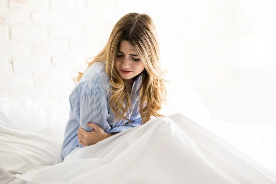 PMS-Therapy A woman with long blonde hair sits in her bed holding her stomach in pain, considering getting care for PMS from Anita Petruzzelli, M.D. in Glastonbury.