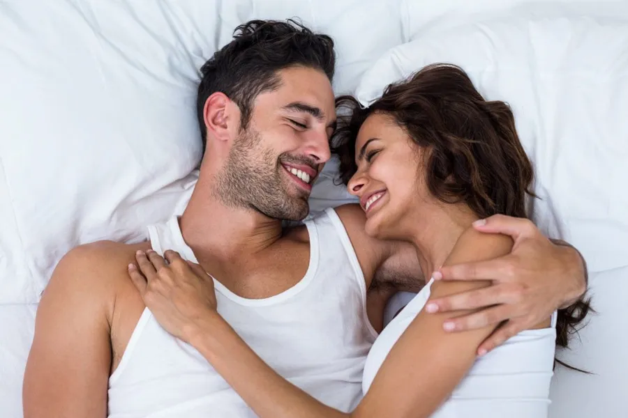 PT141-for-Men-Doctor A laughing man and woman in undershirts lie in each other's arms on a white bed, representing successful PT141 sexual wellness therapy for men from Anita Petruzzelli, M.D. in Glastonbury.