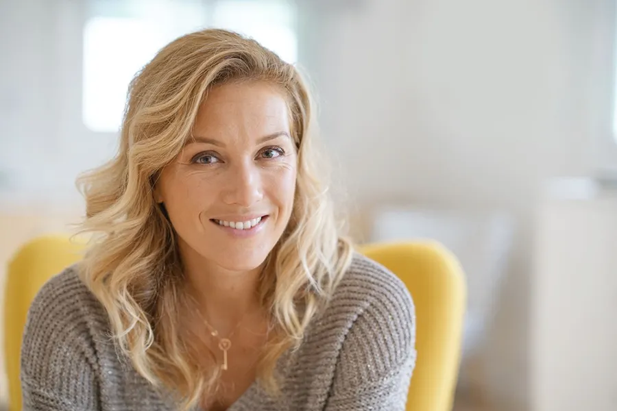Perimenopause-Doctor A blonde woman wearing a a gray sweater sits inside in a yellow chair, smiling and happy with her perimenopause treatment from Anita Petruzzelli, M.D. in Glastonbury.