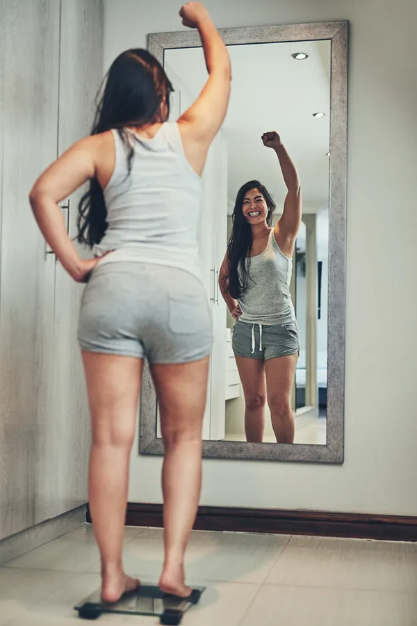 Tirzepatide-Treatment A woman in workout clothes stands on a scale looking in the mirror and pumping her hands in celebration of her successful Tirzepatide treatment from Anita Petruzzelli, M.D. in Glastonbury.